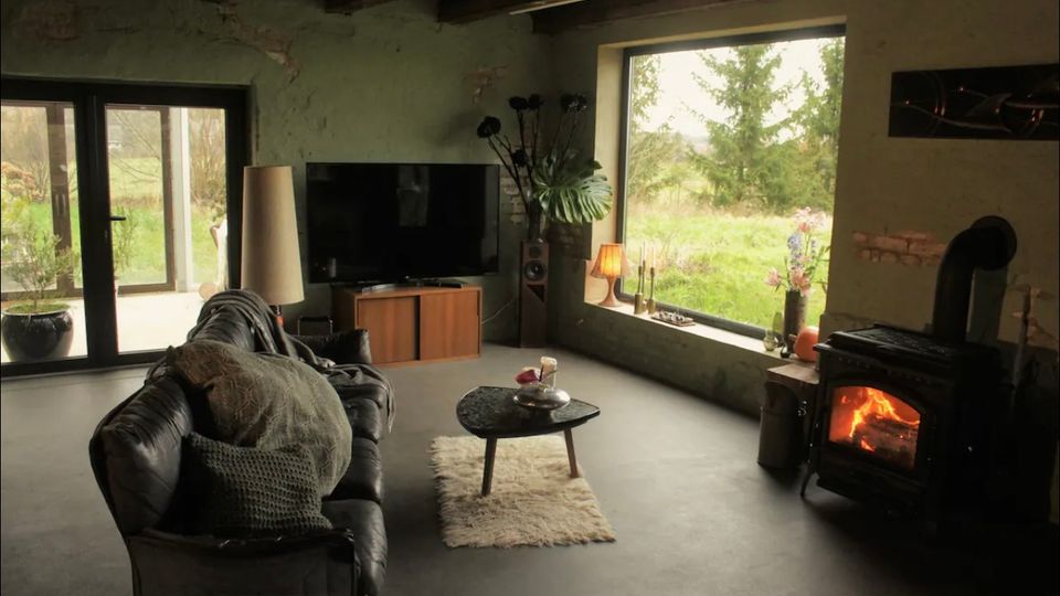 Wohnzimmer in einem rustikalen Stil mit sichtbaren Holzbalken und verputzten Wänden. Im Raum befinden sich ein Kaminofen, eine Ledercouch, ein kleiner Couchtisch auf einem Teppich, sowie ein großes Fenster mit Blick auf eine grüne Landschaft.