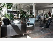 Offener Raum im industriellen Stil mit Sitzbereich, ausgestattet mit Ledersesseln, einem Couchtisch und großen Zimmerpflanzen. Im Hintergrund sind Regale, Schreibtische und weitere Büromöbel sichtbar.