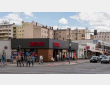 Straßenszene mit einem Rewe-Supermarkt in einem modernen, flachen Gebäude mit dunkler Fassade, umgeben von Wohn- und Geschäftshäusern. Im Vordergrund eine Kreuzung mit Fußgängern und Autos; Schilder und Ladeneingänge von Nachbargeschäften sind sichtbar.