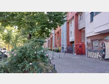 Straßenansicht in einer städtischen Umgebung mit roten Klinkergebäuden und grauen Fassadenelementen, die mit Graffiti bedeckt sind. Im Vordergrund befinden sich blühende Büsche und ein Gehweg mit Kopfsteinpflaster, gesäumt von einer Baumreihe und einer alten Laterne.