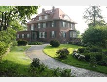 Backsteinhaus im klassischen Stil mit mehreren weißen Sprossenfenstern und grünen Fensterläden, umgeben von einem gepflegten Garten mit blühenden Sträuchern und einem geschwungenen gepflasterten Weg, der zum Eingang führt. Im Hintergrund stehen hohe Bäume, die das Anwesen einfassen.