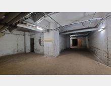 Kellerraum mit rohen Betonwänden, verputzten Pfeilern und sichtbaren Rohrleitungen an der Decke. Der Raum ist schwach beleuchtet durch Deckenlampen, und eine Holztür ist im Hintergrund sichtbar.