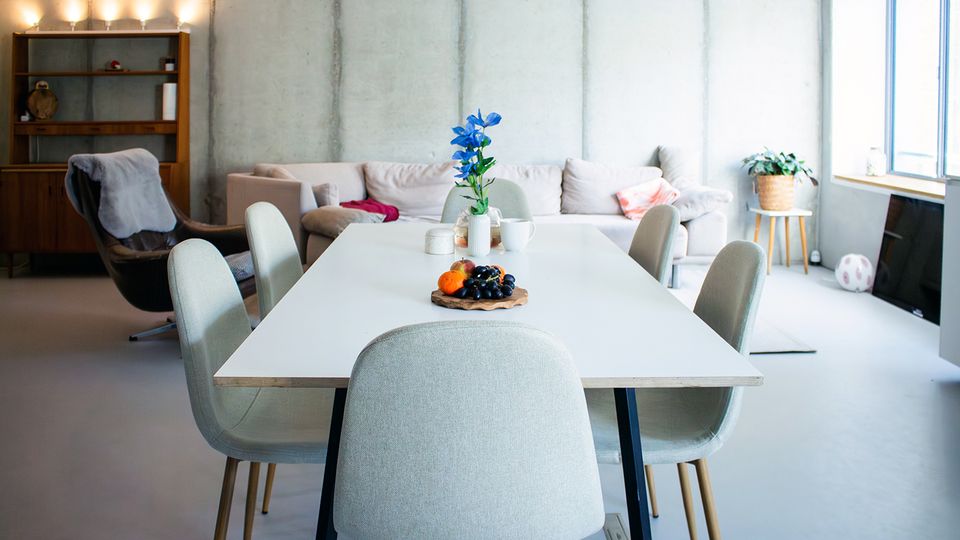 Heller, moderner Ess- und Wohnbereich mit großem weißen Esstisch und gepolsterten Stühlen, dahinter ein Sofa vor einer Sichtbetonwand. Rechts sorgen große Fenster für Tageslicht; auf dem Tisch stehen Blumen und eine Obstschale, links ist ein Regal aus Holz zu sehen.