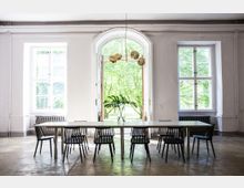 Großer, heller Raum mit hohen Decken und einem langen weißen Tisch, umgeben von schwarzen Stühlen. Drei große Fenster, darunter ein bogenförmiges Mittelfenster, bieten Blick ins Grüne; in der Mitte des Tisches steht eine Vase mit Monstera-Blättern.