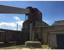 Industriegelände mit alten Ziegel- und Betongebäuden, darunter ein mehrstöckiger Backsteinbau mit großen Fenstern. Im Vordergrund sind eine schmale Metallbrücke, ein Kaminrohr und ein runder Beton- oder Metallsockel zu sehen, umgeben von spärlicher Vegetation.