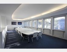 Moderner Besprechungsraum mit langen weißen Tischen, umgeben von weißen Stühlen auf dunkelblauem Teppichboden. Große Fenster auf der rechten Seite lassen viel Tageslicht herein, und an der Rückwand hängen gerahmte Bilder.