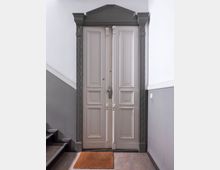 Doppelflügeltür aus Holz mit verzierten Kassetten und einem dunkelgrauen, dekorativen Türrahmen mit Ornamenten. Im Vordergrund liegt eine braune Fußmatte, seitlich sind eine Treppe und eine weiß-graue Wand sichtbar.