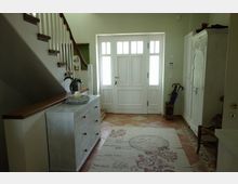 Heller Eingangsbereich eines Hauses mit weißer Eingangstür und Glasfenstern, gefliesten Boden in warmen Tönen und einer dekorativen Kommode links unter einer Holztreppe. Rechts steht ein weißer Kleiderschrank, daneben ein Ständer mit Regenschirmen.