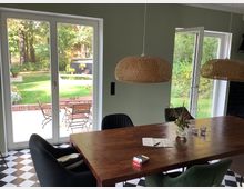 Esszimmer mit großem Holztisch, schwarzen und grünen Stühlen, geflochtenen Hängelampen und schwarz-weiß kariertem Boden. Im Hintergrund führen bodentiefe Fenster und eine geöffnete Tür zu einer Terrasse mit Gartenmöbeln und einem gepflegten Rasen.