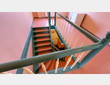 Treppenhaus mit grüner, geländerumrahmter Treppe und cremefarbenen Balustern in einem Gebäude mit rosa gestrichenen Wänden und orangefarbenem Bodenbelag. Im Hintergrund ein Heizkörper und ein weißer Schrank, die an der Wand stehen.