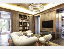 Wohnzimmer in einem gehobenen Stil mit einer cremefarbenen, gepolsterten Chaiselongue, zwei Stühlen mit weißem Fellbezug, einem runden Couchtisch und einem großen Holzregal mit Büchern und Dekoration. An der Decke hängt ein großer, glitzernder Kronleuchter, und ein breites Fenster im Hintergrund öffnet den Raum zum Außenbereich.