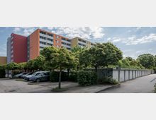 Wohnanlage mit mehrstöckigen, farbigen Wohngebäuden im Hintergrund, davor eine Reihe von kleinen Bäumen, geparkten Autos und Garagen. Der Bereich ist umgeben von Gehwegen und Grünflächen unter einem bewölkten Himmel.