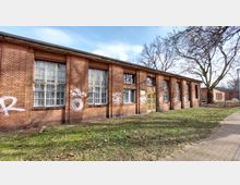 Backsteingebäude mit großen Fenstern und gelber Eingangstür, teilweise mit Graffiti besprüht. Im Vordergrund eine Grünfläche und ein gepflasterter Gehweg, im Hintergrund kahle Bäume.