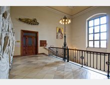 Ein historischer Innenraum mit einer breiten Treppenabsatzfläche, flankiert von einer Brüstung mit kunstvoll verziertem Metallgeländer. An der Wand befinden sich eine Holztür mit Glaseinsätzen, Reliefskulpturen, ein Gemälde sowie ein großes Fenster, das Tageslicht hereinlässt.