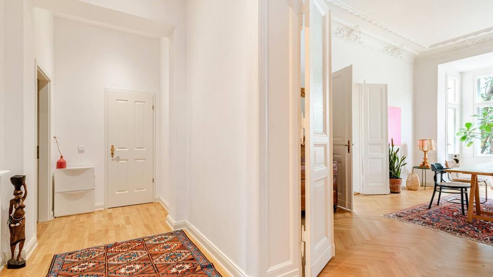 Heller Flur in einer Altbauwohnung mit weißen Wänden, Stuckdecke, hohen Türen und Parkettboden mit gemusterten Teppichen. Rechts öffnet sich der Blick in ein Zimmer mit großen Fenstern, Tisch und Stühlen sowie Zimmerpflanzen.