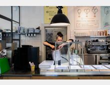 Eisdiele mit moderner Einrichtung: Eine Mitarbeiterin hinter dem Tresen füllt Eis aus einem Behälter in die Kühlvitrine. Im Hintergrund sind eine Kaffeemaschine, Regale mit Geschirr und eine Menütafel an der Wand sichtbar.