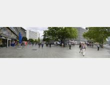 Weitläufiger Platz in einer urbanen Umgebung mit Gehwegplatten, umgeben von modernen Gebäuden. Im Vordergrund sind Passanten und Verkaufsstände, im Hintergrund stehen Bäume und ein Gebäude mit einer auffälligen Gitterstruktur.