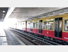 Bahnsteig an einer überdachten S-Bahn-Station mit einer vorbeifahrenden rot-gelben S-Bahn. Im Hintergrund ist ein heller Ausgang sichtbar, der von Tageslicht erleuchtet wird.