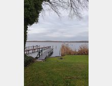 Ein Seeufer mit einem schmalen Holzsteg, der ins Wasser führt. Im Vordergrund ist eine grüne Rasenfläche mit einem Feuerstellenring, rechts davon stehen Schilfpflanzen, und links ragt ein Baum mit kargen Ästen ins Bild.