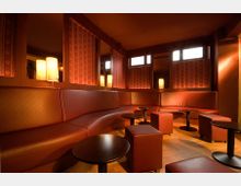 Ein gemütlicher Lounge-Bereich mit geschwungener, gepolsterter Sitzbank in braunem Leder, gepaart mit kleinen runden Tischen und würfelförmigen Hockern. Die Wandpaneele sind mit dekorativen Mustern gestaltet, während warme Lampen und kleine Fenster die Beleuchtung ergänzen.