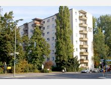 Mehrstöckiges Wohnhaus mit beigen Fassaden, Balkonen und vereinzelter Begrünung, umgeben von hohen Bäumen. Im Vordergrund befinden sich eine Straße mit geparkten Autos, eine Fußgängerampel und eine Person mit Kinderwagen an einer Straßenecke.