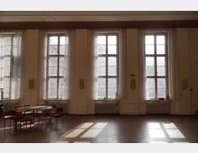 Großer Raum mit hohen Decken und vier großen Fenstern mit Sprossen, durch die Tageslicht einfällt. Im Vordergrund stehen ein Tisch und mehrere Stühle, der Boden ist glatt und der Raum wirkt hell und schlicht.