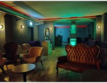 Ein stilvoll eingerichteter Lounge-Bereich mit retro-inspirierten Sesseln und einem lederbezogenen Sofa auf einem Holzboden. Die Decke ist mit farbiger LED-Beleuchtung und sichtbaren Lüftungsrohren gestaltet, während sanfte Wandlampen für stimmungsvolles Licht sorgen.