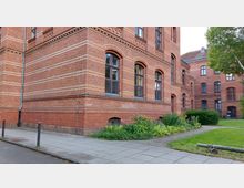 Backsteinbau im Stil des Historismus mit hohen Fenstern und dekorativen Verzierungen. Im Vordergrund ein gepflasterter Bürgersteig, ein kleines Blumenbeet und eine Rasenfläche mit einem großen Baum.