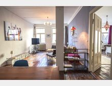 Wohnzimmer in einer Wohnung mit Holzboden und weiß gestrichenen Wänden. Der Raum ist eingerichtet mit einem Fernseher, Sofas, einem Bücherregal und dekorativen Lampen, während im Hintergrund durch eine offene Tür ein weiterer Raum mit einem roten Kronleuchter sichtbar ist.