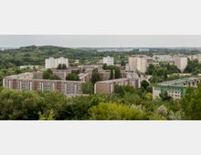 Wohngebiet mit mehrstöckigen Plattenbaugebäuden, umgeben von dichtem Grün und Bäumen. Im Hintergrund eine hügelige Landschaft und weitere Wohngebäude, die sich bis zum Horizont erstrecken.