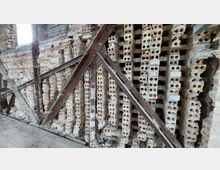 Ziegelwand in einem alten, teils verfallenen Gebäude, verstärkt durch diagonale Metallstreben. Die Ziegel sind unregelmäßig gestapelt, mit sichtbaren Hohlräumen und stellen den Eindruck eines nicht vollständig fertigen oder stabilen Bauwerks dar.