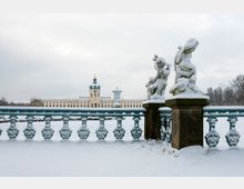 Schneebedeckter Garten mit Blick auf ein barockes Schloss im Hintergrund, das eine zentrale Kuppel und helle gelbe Fassaden aufweist. Im Vordergrund stehen verzierte Steinfiguren und ein mit Schnee bedecktes schmiedeeisernes Geländer.
