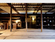 Ein rustikaler Dachboden mit Holzboden und freigelegten Holzsäulen, umgeben von Regalen mit Haushaltsgegenständen und Möbeln. Der Raum ist spärlich beleuchtet durch mehrere Tischlampen, während die Decke aus Metallplatten besteht.