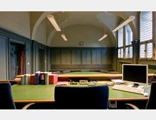 Innenaufnahme eines Gerichtssaals mit hoher Decke und gewölbtem Bogen. Im Vordergrund steht ein großer Tisch mit grüner Arbeitsfläche, Büchern, einem Computer und Büroutensilien, während der Hintergrund durch Holzvertäfelungen, eine Wanduhr und hohe Fenster mit Gitterglas geprägt ist.