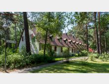 Mehrfamilienhaus im traditionellen Stil mit weißer Fassade, roten Fensterläden und einem Dach mit mehreren Dachgauben, umgeben von hohen Bäumen und dichten Sträuchern. Links im Bild befindet sich ein Straßenlaterne samt Wegweiser mit der Aufschrift „Jugenheimer Weg“.