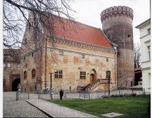 Historisches Gebäude aus Backstein mit einem roten Satteldach und einer runden, steinernen Turmstruktur an der Seite. Der Hof ist mit Kopfsteinpflaster und einer kleinen Grünfläche gestaltet, umgeben von einem Metallzaun.