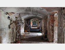 Ein verlassener, schmaler Korridor mit gewölbter Decke, bröckelnden Wänden aus Ziegelstein und Putz sowie heruntergekommenem Boden aus Ziegelsteinen. An den Wänden hängen vertrocknete Äste, Stofffetzen und weitere Objekte; im Hintergrund fällt Licht durch eine Türöffnung.