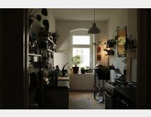 Schmale Wohnküche mit hohen weißen Wänden und großem Fenster am Ende, dessen Fensterbank mit Topfpflanzen und Kräutern bestückt ist. Links und rechts stehen dunkle Arbeitsflächen, offene Regale mit Gläsern und Küchenutensilien sowie ein Gasherd; eine Hängelampe und Wanddekorationen ergänzen den Raum.