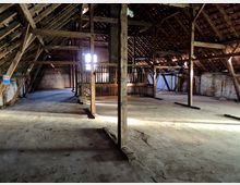 Offener Dachbodenraum eines alten Gebäudes mit hölzernen Dachbalken, schrägem Ziegeldach und teilweise verputzten Backsteinwänden. Der Boden ist aus Beton, und die Sonnenstrahlen fallen durch Lücken im Dach ein.