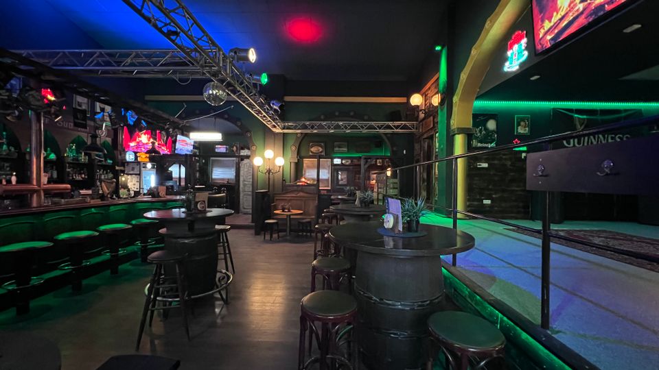 Innenraum einer Irish-Pub-ähnlichen Bar mit langer Theke, hohen Rundtischen und Barhockern sowie dunkler Holz- und Metalloptik. Neon- und Bühnenbeleuchtung mit Traversen an der Decke, mehrere Bildschirme und ein leicht erhöhter Bereich mit Geländer an der rechten Seite.