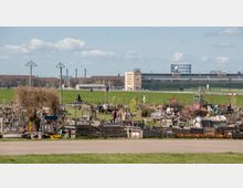 Städtischer Gemeinschaftsgarten mit Beeten aus Holzpaletten und improvisierten Strukturen auf einer offenen Wiese, im Hintergrund das ehemalige Flughafen-Gebäude Tempelhof und ein großes, freies Feld.