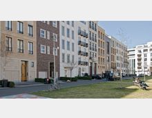 Reihe von modernen städtischen Wohngebäuden mit unterschiedlichen Fassadenmaterialien, darunter Backstein und Putz, entlang einer gepflasterten Straße. Im Vordergrund befinden sich eine Grünfläche mit Bäumen, Sitzbänken und parkenden Autos.