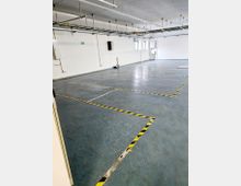 Leerer Innenraum mit grauem Boden, gelb-schwarzen Markierungen auf dem Boden und weißen Wänden. An der Decke befinden sich Kabeltrassen und Beleuchtung, an den Wänden sind elektrische Anschlüsse und Notausgangsschilder sichtbar.