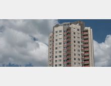 Wohnhochhaus mit mehreren Etagen und Balkonen, gestaltet in grauem Beton mit roten Akzentlinien, vor einem wolkigen Himmel.