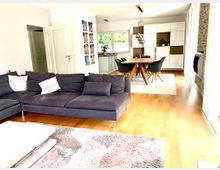 Helles Wohnzimmer und Esszimmer in einem offenen Grundriss, mit einem großen grauen Ecksofa, Holzboden, einem rechteckigen Esstisch mit vier Stühlen und einer modernen, weißen Einbauküche im Hintergrund. Große Fenster lassen Tageslicht herein, und eine Steinwand mit einem eingebauten Kamin ist auf der rechten Seite sichtbar.