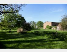 Weite, grüne Wiese mit einigen Bäumen und Sträuchern, im Hintergrund ein großes Gebäude aus rotem Ziegelstein. Der Himmel ist klar und blau, die Szene liegt in ländlicher Umgebung.