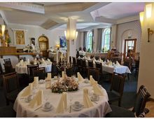 Ein festlich dekorierter Speisesaal mit runden und rechteckigen Tischen, die mit weißen Tischdecken, gefalteten Stoffservietten, Blumengestecken und Kerzenständern gedeckt sind. Die Wände sind mit Wandverzierungen, großen Fenstern und klassischem Dekor gestaltet, während der Raum eine helle und elegante Atmosphäre vermittelt.