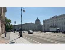 Straßenansicht in einer Stadt mit einem breiten Gehweg, Laternen, vorbeifahrenden Autos und Fußgängern. Im Hintergrund ist ein großes historisches Gebäude mit einer Kuppel zu sehen, umgeben von weiteren Gebäuden im klassischen Architekturstil.