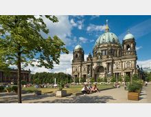 Ansicht des Berliner Doms, einer historischen Kirche mit grünen Kuppeln und reich verzierten Fassaden, umgeben von Grünanlagen und einem Platz, auf dem Menschen entspannen. Im Vordergrund stehen ein Baum und Pflanzkästen, während der Himmel klar und blau ist.