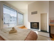 Helles Schlafzimmer mit großen Fenstern, die durch Jalousien teilweise abgeschirmt sind. Im Raum befinden sich ein gemachtes Bett mit gefalteten Handtüchern und ein moderner Kamin in einer weißen Wand integriert.